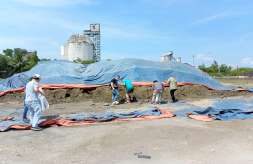 Workers placing tarpaulins over mineral sand stockpile with industrial silos in the background